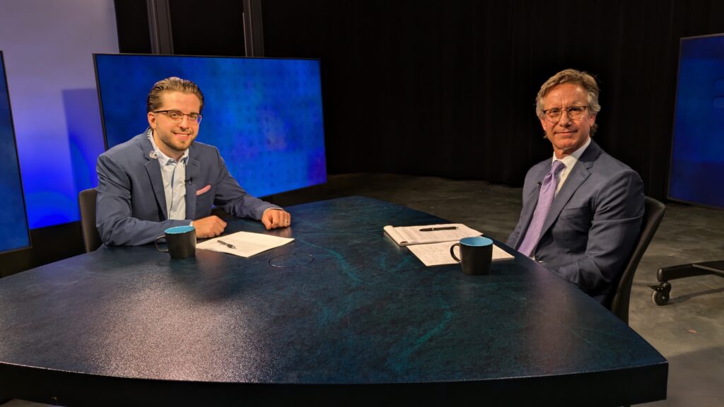 Two men in suits are seated at a blue desk with papers and black coffee mugs in a studio. They appear poised for an interview or discussion.