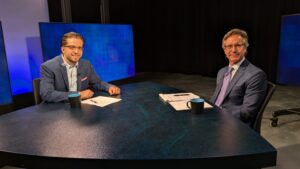 Two men in suits are seated at a blue desk with papers and black coffee mugs in a studio. They appear poised for an interview or discussion.