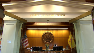 Entrance to the Office of the Governor, featuring a seal on the wooden wall, two flags on either side, and glass doors.