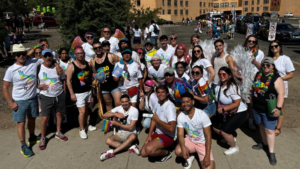 A group of people in colorful attire and accessories gather together outdoors, posing for a photo during a public event. They are holding up flags and wearing shirts with a vibrant design.
