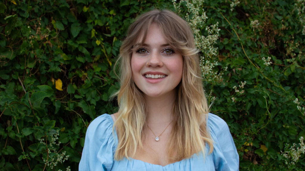 A young woman with long, blonde hair is smiling and standing in front of green foliage wearing a light blue blouse.