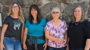 Four women standing in front of a stone wall, smiling, with casual attire including glasses, t-shirts, and jeans. The cool blue of their outfits hints at the calmness of water.