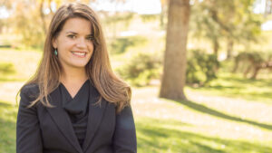 A woman with long hair wearing a black blazer is standing outside in a sunlit park with trees and grass in the background.