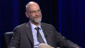 A bald man with glasses, a gray beard, and a suit is seated and smiling against a dark blue backdrop.