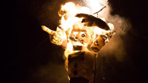A large puppet is set on fire at night, showing flames and smoke as it burns.