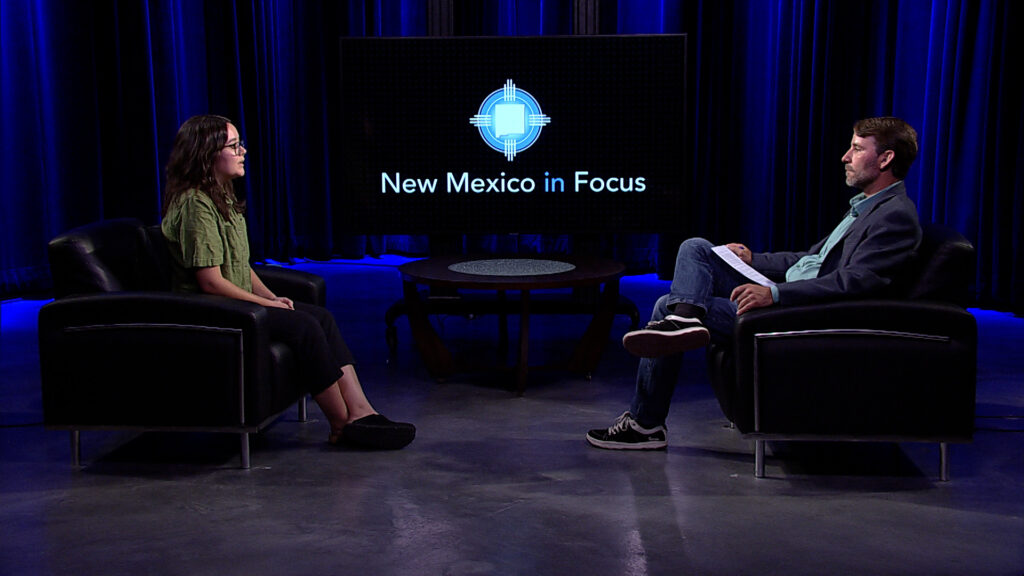 Two people sit across from each other in a studio with a "New Mexico in Focus" sign displayed on a large screen behind them, engaging in a conversation. The studio has blue lighting and dark curtains.