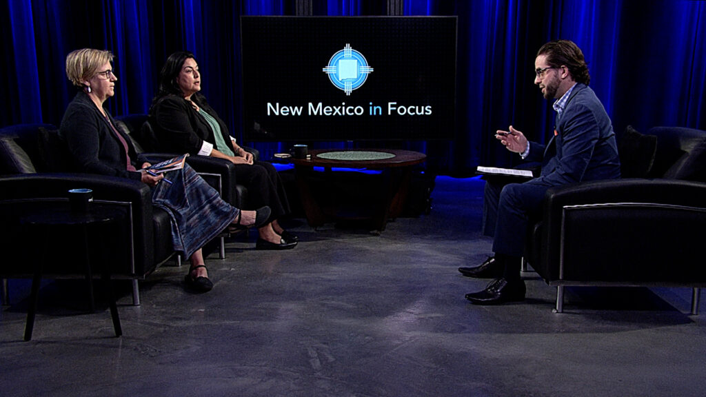 Three individuals engaged in a discussion on a television set with a large screen displaying "New Mexico in Focus" in the background. They are seated in a studio setting with blue curtains.
