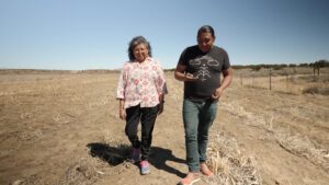 Two people walking and conversing in a dry, open field under a clear sky. The woman on the left wears a patterned blouse, and the man on the right wears a T-shirt and jeans while looking at his phone, perhaps searching for nearby water sources.