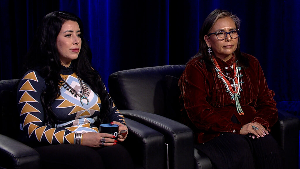 Two women sit on black leather chairs against a dark backdrop. The woman on the left wears a geometric-patterned top, and the woman on the right wears a red shirt with layered necklaces.