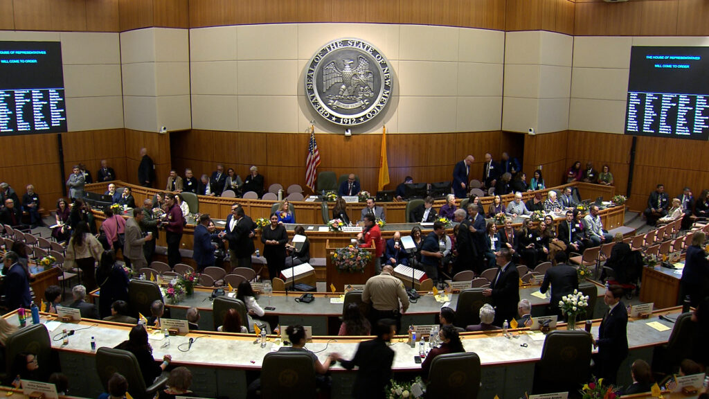 People gather in a legislative chamber with a large seal on the wall, digital boards displaying information, and floral arrangements on desks.