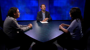 Three people sit around a triangular table on a TV set with blue background screens. The person in the center has a paper in front of them, appearing to moderate a discussion.