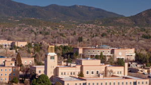 Aerial view of a beige building complex with a clock tower, surrounded by trees and backed by mountains under a clear blue sky.