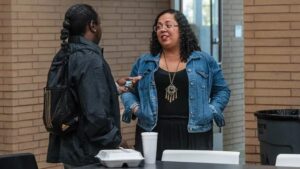 Two people stand and talk indoors near a table with food containers and a cup. One person holds a phone while the other listens. Behind them is a brick wall.