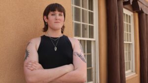 Person with a black tank top and tattoos stands with arms crossed against a beige wall next to a window.