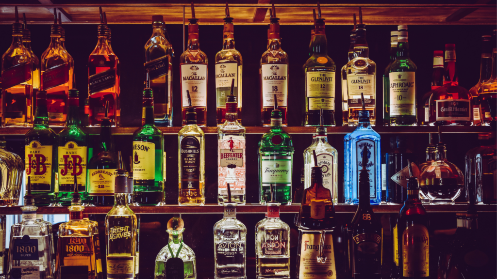 A variety of liquor bottles are displayed on wooden shelves, featuring brands of whiskey, gin, rum, and tequila. The background is dimly lit, highlighting the bottles' labels.