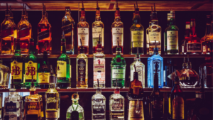 A variety of liquor bottles are displayed on wooden shelves, featuring brands of whiskey, gin, rum, and tequila. The background is dimly lit, highlighting the bottles' labels.