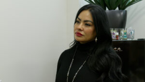 Person with long black hair and a black outfit stands in a room with a plant and small items on a shelf in the background.