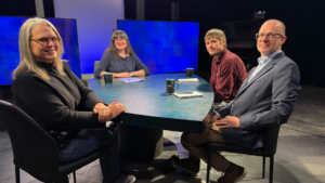 Four people sit at a rounded rectangular table in a studio setting with black mugs in front of them.