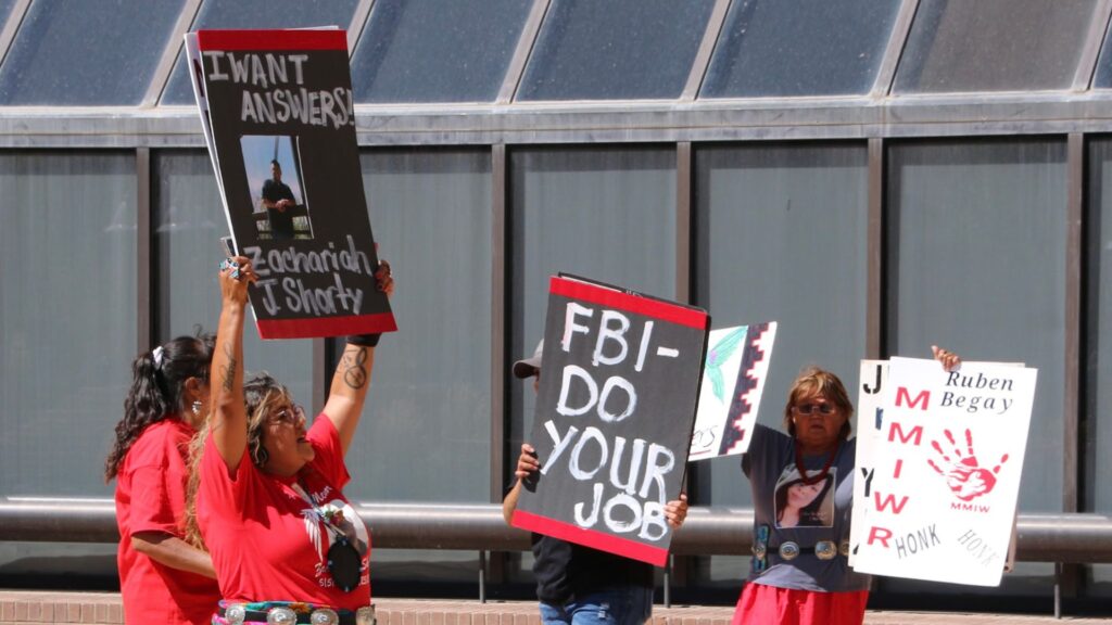 Protesters hold signs demanding action and answers. One reads "I want answers" with a photo, another "FBI - Do Your Job," and a third mentions Ruben Begay and justice.