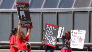Protesters hold signs demanding action and answers. One reads "I want answers" with a photo, another "FBI - Do Your Job," and a third mentions Ruben Begay and justice.