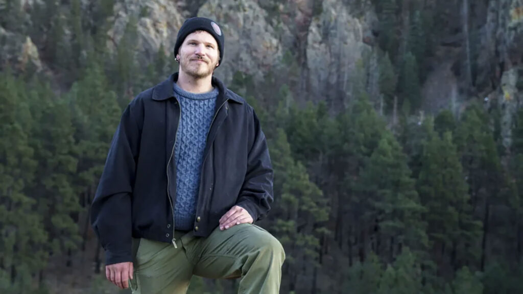 A man wearing a black jacket, blue sweater, and beanie poses with one knee up in front of a forested mountain landscape.