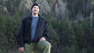A man wearing a black jacket, blue sweater, and beanie poses with one knee up in front of a forested mountain landscape.