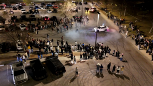 Aerial view of a large crowd watching a car performing stunts in a parking lot at night, surrounded by parked vehicles. Smoke is visible from the car's tires.