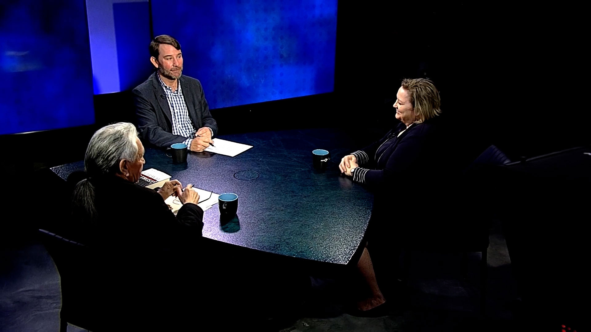 Three people sit around a round table with papers and coffee mugs, engaged in a discussion in a studio with blue lighting.
