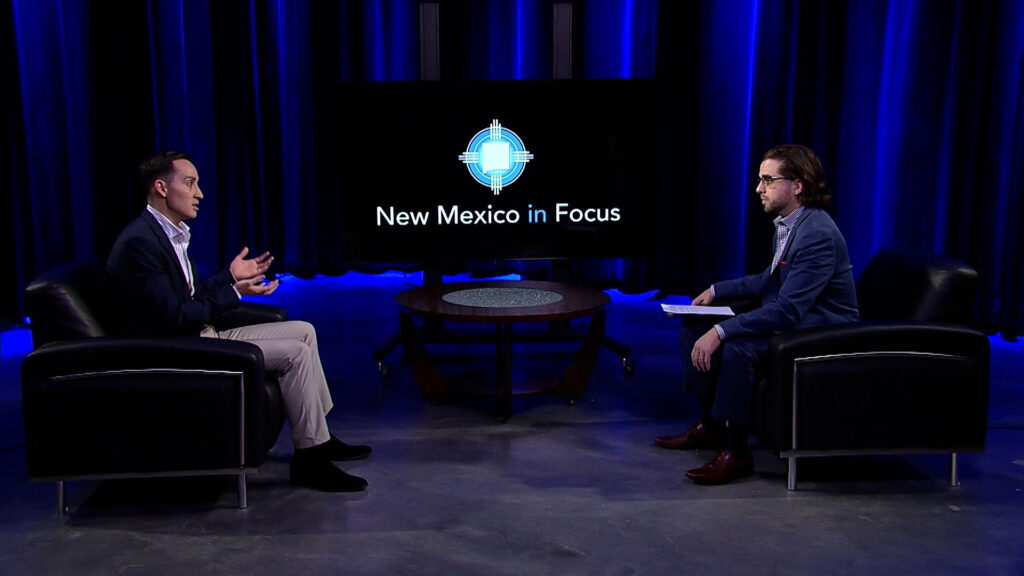 Two men in suits sit across from each other in a TV studio with a "New Mexico in Focus" logo displayed on a screen between them.