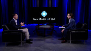 Two men in suits sit across from each other in a TV studio with a "New Mexico in Focus" logo displayed on a screen between them.
