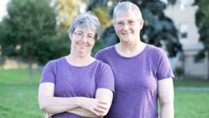 Two people with short gray hair, glasses, and matching purple shirts stand together outdoors, smiling at the camera. Trees and a building are visible in the background.