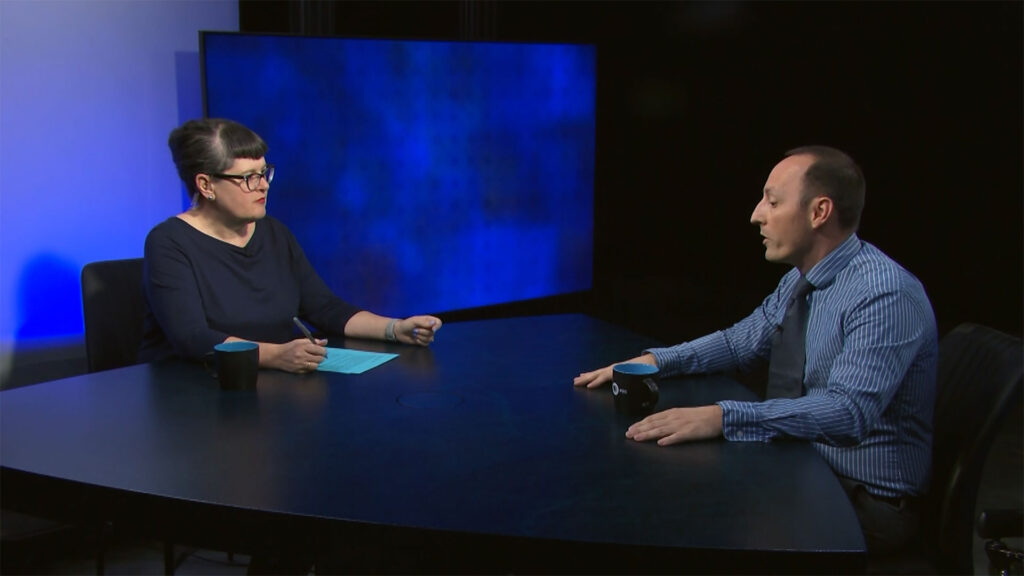 Two people are seated at a round table having a discussion; the woman holds a pen and paper while the man gestures with his hands. A large blue screen is visible in the background.