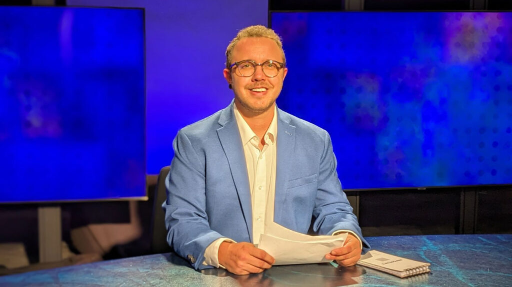 A person in a blue suit jacket and glasses sits at a desk with papers, in front of blue television screens in a studio setting.