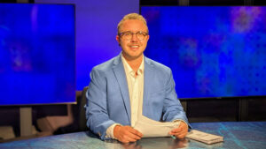 A person in a blue suit jacket and glasses sits at a desk with papers, in front of blue television screens in a studio setting.