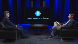 Two men sit facing each other in a studio with a large screen displaying "New Mexico in Focus" between them. Both are seated on black chairs against a backdrop of blue curtains.