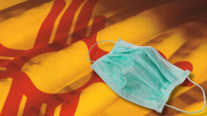 A blue surgical mask rests on a New Mexico state flag with red and yellow colors and the Zia sun symbol.