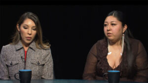 Two women sit side by side at a table with two blue mugs, facing forward against a dark background.