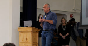 An older man in a denim shirt and jeans is speaking into a microphone at a wooden podium. A woman and others stand in the background.