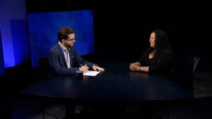 A man in a suit interviews a woman with curly hair at a dark table in a studio with blue-lit backgrounds.