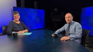 Two people sit at a round table with coffee mugs and notes, facing the camera in a television studio with blue screens and studio equipment in the background.