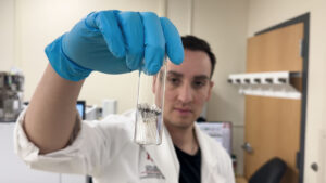 A scientist in a lab coat and blue gloves holds up a small glass vial containing metal samples in a laboratory setting.