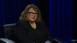 A woman with glasses, wearing a black outfit, is seated against a dark blue curtain backdrop.