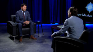 Two men in suits sit across from each other in a studio with black chairs, blue lighting, and a "New Mexico" logo visible in the background.