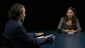 Two people are sitting at a round table in a studio setting, engaged in conversation. One person is writing in a notebook, and there are cups on the table.