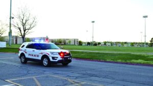 A police SUV with flashing lights is parked on a road near a grassy field, on a clear day, with sports field lights and school buildings in the background.