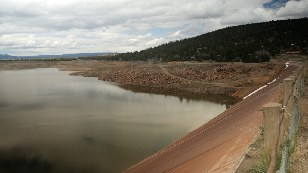 A reservoir with low water levels bordered by rocky terrain and a hillside under a cloudy sky.