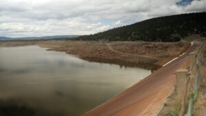 A reservoir with low water levels bordered by rocky terrain and a hillside under a cloudy sky.