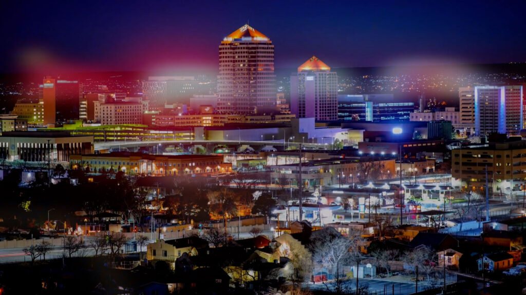 A cityscape at night shows illuminated buildings, including two high-rise towers with orange-lit roofs, against a dark sky with city lights in the background.