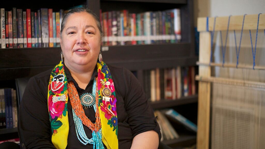 A woman wearing colorful jewelry and a floral scarf sits in front of bookshelves and a wooden loom.