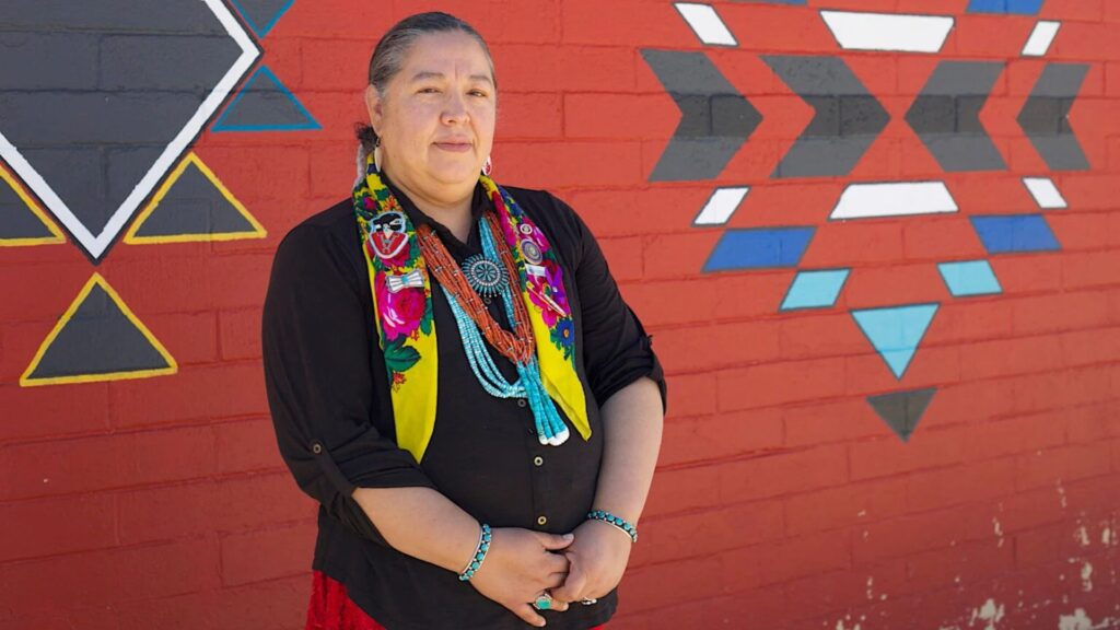 A person with long hair in a braid stands in front of a red brick wall with geometric patterns, wearing colorful jewelry and a floral scarf.